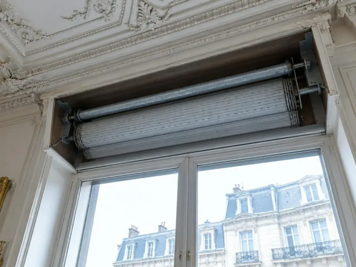 Coffre intérieur ouvert sous un plafond mouluré, axe métallique et tablier enroulé au-dessus d'une fenêtre